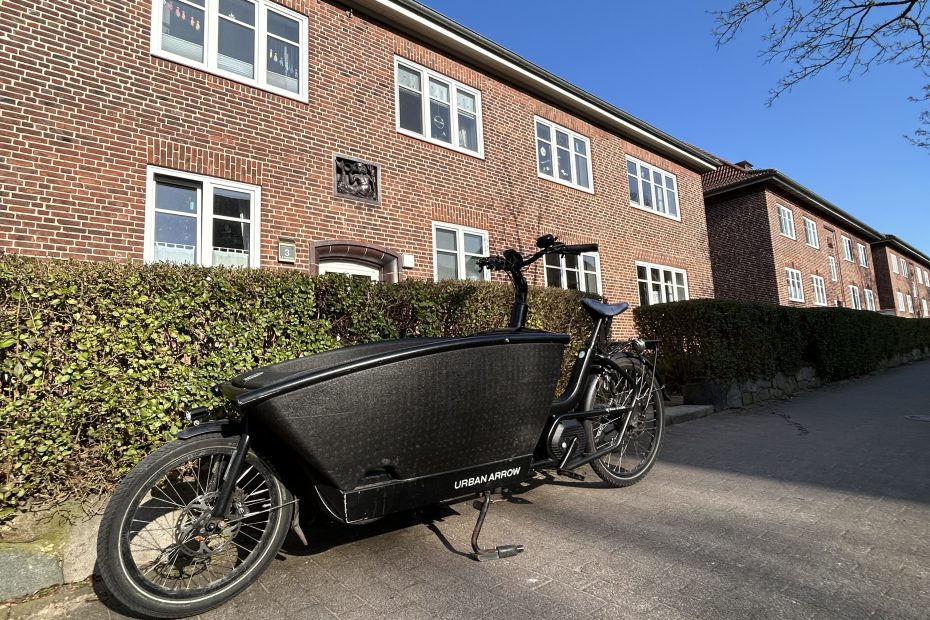 Schwarzes Lastenfahrrad vor der Vorgarten-Hecke eines Mehrfamilienhauses abgestellt